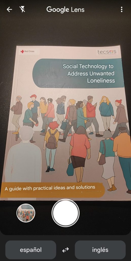 Se muestra la cámara "Google Lens" enfocando la guía para la soledad no deseada, cuyo título se muestra traducido al inglés "Social technology to address unwanted loneliness". En la parte inferior central se encuentra el botón de la cámara para realizar la foto. En la parte superior izquierda aparece el botón para retroceder y, junto a este, el del flash de la cámara.